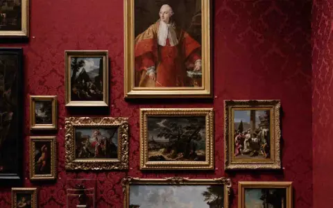 A gallery wall displays classical paintings in ornate frames against a rich red damask background. This collection showcases the best way to invest in art, with portraits and landscapes hinting at historical narratives.
