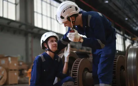 Kierownik logistyki w białym kasku i niebieskim kombinezonie rozmawia przez krótkofalówkę z koleżanką, omawiając szczegóły dostawy.