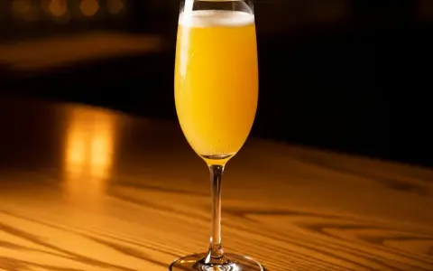Ett glas med en gyllene mimosa drink, bubblig och uppfriskande, står på ett träbord i varmt ljus.