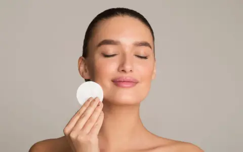 Donna con occhi chiusi applica un dischetto di cotone sul viso, parte della sua routine di skincare viso per una pelle radiosa.