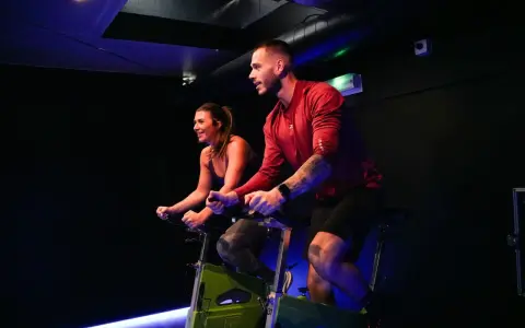 Un homme et une femme s'entraînent sur des vélos d'appartement dans une salle de sport sombre, profitant d'une séance de rpm sport intense.