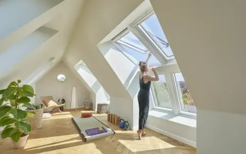 Frau öffnet Dachfenster in hellem Dachboden-Yoga-Raum. Hier darf man Dachboden als Wohnraum nutzen und schafft sich eine Oase.