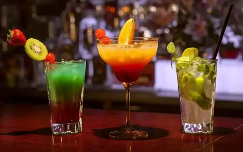 Trzy kolorowe drinki na barze. Jeden z nich to drink z pomarańczą, ozdobiony owocami.