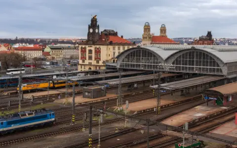 Widok na dworzec kolejowy w Pradze z pociągami na torach i budynkami w tle.