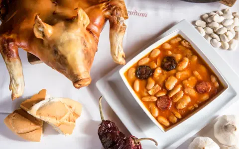 Cochinillo asado segoviano, crujiente y dorado, acompañado de un plato de judiones con chorizo y morcilla, pan y ajos.