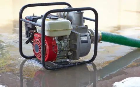 Una motobomba roja y plateada, con una manguera verde, está sumergida en agua turbia, lista para drenar.