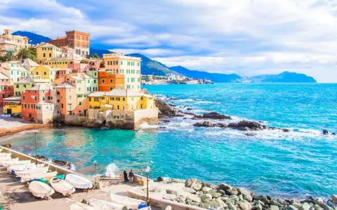Case colorate a picco sul mare turchese, barche ormeggiate e un promontorio roccioso: un angolo di Genova che ricorda le sue spiagge più belle.