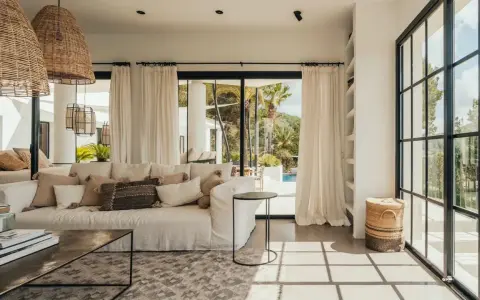 Arredare una casa al mare: divano bianco, cuscini naturali, lampade in rattan e vista su piscina e palme.