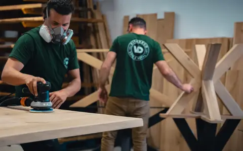 Un artisan masqué utilise une ponceuse pour poncer une table en bois. Un autre artisan travaille sur un pied de table en bois en arrière-plan.
