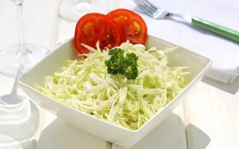 Frischer, selbstgemachter Krautsalat in einer weißen Schüssel, garniert mit Petersilie und Tomatenscheiben. Daneben liegen ein grüner Löffel und eine Serviette.