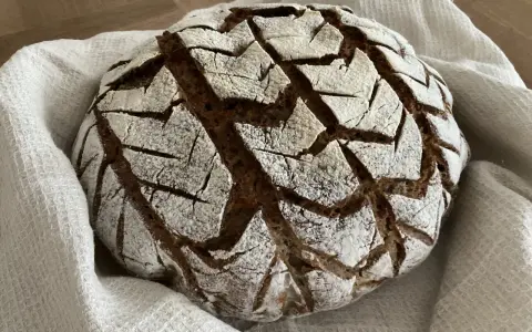 Ein rustikales, rundes buchweizenbrot sauerteig mit einer gemusterten Kruste, bestäubt mit Mehl, liegt auf einem weißen Leinentuch.