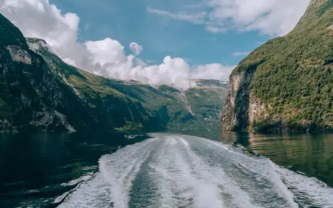 Rejs po Geirangerfjordzie. Pogoda sprzyja, chmury wiszą nad zielonymi zboczami gór, a za statkiem widać biały ślad na spokojnej wodzie.
