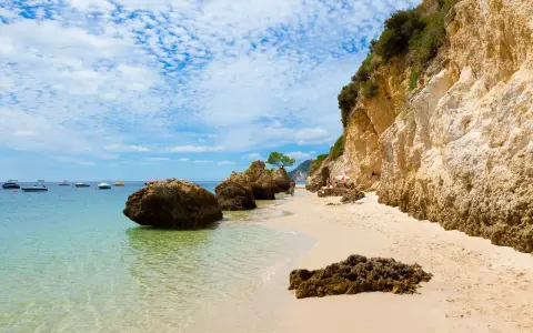 Sonniger Setúbal Strand mit türkisfarbenem Wasser, Felsen und Booten im Hintergrund. Eine kleine Gruppe entspannt sich unter einem Sonnenschirm.