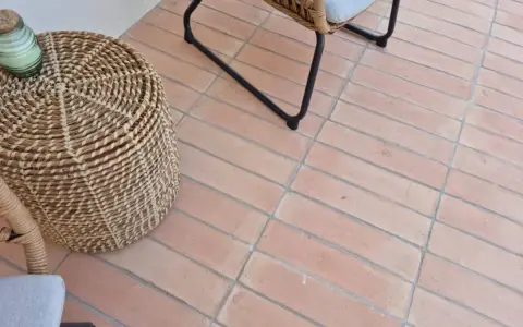 Suelo de baldosas de terracota con juntas limpias, listo para rellenar juntas baldosas suelo. Un cesto de mimbre y una silla de ratán completan la escena.