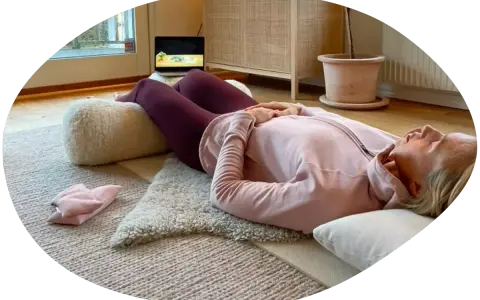 Kvinna ligger på rygg på en matta med en kudde under knäna, hon försöker varva ner med en laptop i bakgrunden.