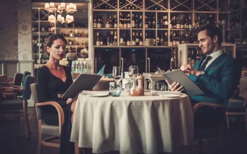 Elegancka para przegląda menu w luksusowej restauracji. On w granatowym garniturze, ona w czarnej sukni.