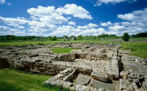 Extensive roman ruin spread across a green field under a blue sky with fluffy clouds.