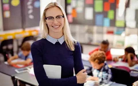 Uśmiechnięta nauczycielka w okularach z tabletem w ręku, dzieci uczą się w tle. To inspiracja, jak zostać nauczycielem.