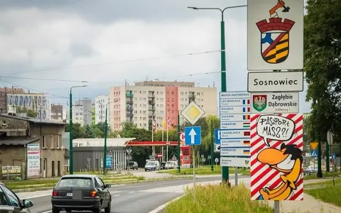 Znak drogowy "Sosnowiec" i humorystyczny napis "Paszport masz?!" przy wjeździe do miasta. W tle bloki mieszkalne.