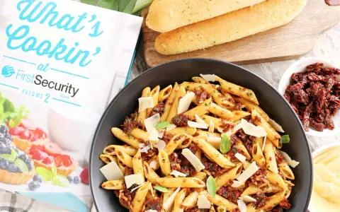 Makaron penne z suszonymi pomidorami, parmezanem i bazylią w czarnej misce, obok pieczywa i książki kucharskiej.