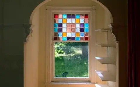Un vitrail design aux carrés colorés illumine une fenêtre, projetant des reflets joyeux sur l'herbe verdoyante à l'extérieur.