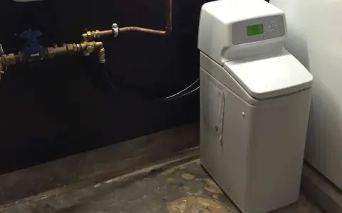 Appareil blanc avec écran vert, connecté à des tuyaux en cuivre. C'est un adoucisseur d'eau potable pour une maison.