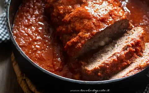 Un succulento polpettone al forno, affogato in un sugo rosso intenso, con una fetta tagliata che rivela un cuore filante di formaggio.