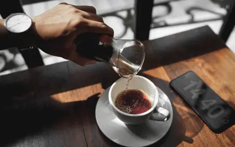 Hand gießt Kaffee in eine Tasse, daneben ein Smartphone mit Uhrzeit.