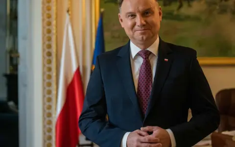 Prezydent Andrzej Duda w garniturze, z polską flagą w tle, czeka na moment, kiedy prezydent podpisze emerytury stażowe.