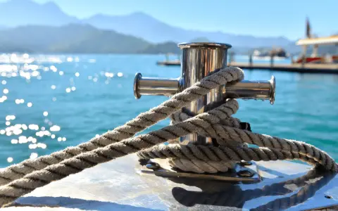 Gros plan sur une ligne de mouillage corps mort, une corde épaisse enroulée autour d'un taquet en acier inoxydable sur un bateau. L'eau bleue scintillante et les montagnes floues en arrière-plan créent une scène paisible.
