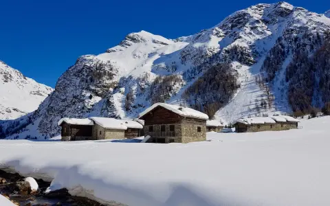 Case innevate nel Val di Rezzalo, con baite in pietra e legno. Per sapere val di rezzalo come arrivare, segui il ruscello che scorre tra la neve.