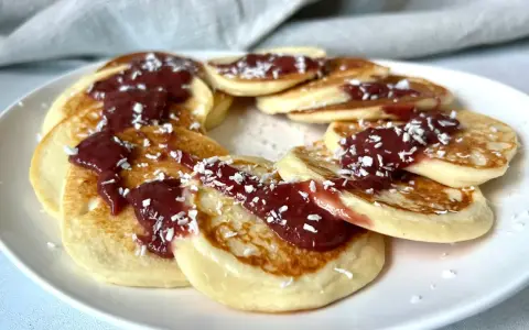 Placki serowe keto ułożone w okrąg na białym talerzu, polane konfiturą i posypane wiórkami kokosowymi.