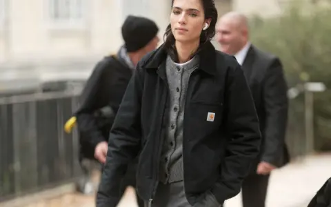 Donna con giacca Carhartt nera e cardigan grigio, cuffiette bianche, cammina con sicurezza.