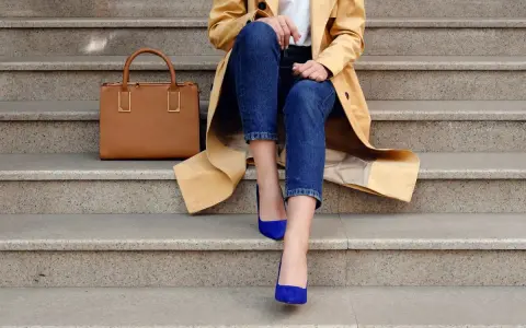 Elegancka stylizacja: beżowy płaszcz, jeansy i buty w kolorze habrowym. Obok torebka w odcieniu karmelu.