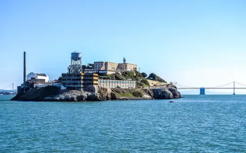 Słynne więzienie Alcatraz na skalistej wyspie, z charakterystyczną wieżą ciśnień i kominem, na tle błękitnego nieba i mostu.