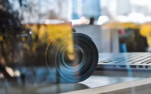 Winieta fotografia: obiektyw Canon na tle laptopa, odbicia tworzą artystyczny efekt.