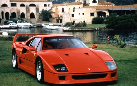 Roter Ferrari F40 auf einer Wiese, im Hintergrund ein Hafen mit Booten und Häusern. Der Ferrari F40 Neupreis war damals ein Traum.