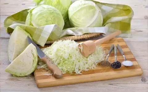 Przygotowanie kiszonej kapusty: ile soli na 10kg? Na desce poszatkowana kapusta, nóż, tłuczek, kminek, pieprz i sól.