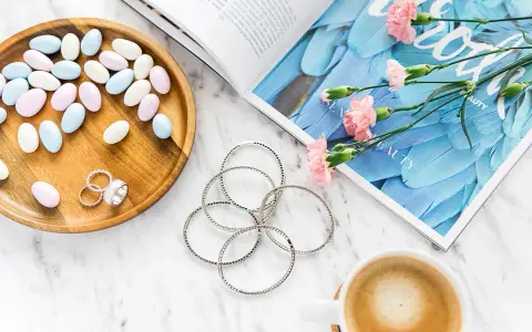Uroczy flat lay: pastelowe drażetki, srebrna biżuteria, różowe goździki i kawa na marmurowym blacie.