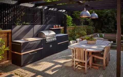 Nowoczesny grill w ogrodzie, z czarną zabudową i drewnianym stołem, zaprasza do wspólnego biesiadowania.