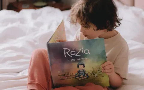Dziecko czyta książkę "Rózia i jej wartości". Obok leżą karty pracy do nauki dodawania i odejmowania pełnych dziesiątek.