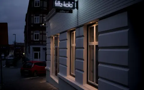 Neon-Schild "fritz-kola" über dem Eingang "dritter ort kiel". Ein rotes Auto parkt auf der Straße.