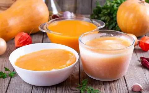 Pomarańczowe zupy w miskach i szklance, dynie i warzywa na drewnianym stole.