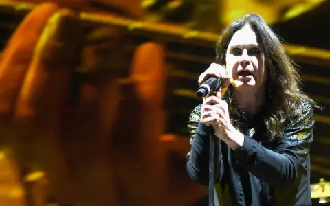 Ozzy Osbourne śpiewa do mikrofonu na koncercie, z rozmytym tłem w kolorach żółci i pomarańczy.
