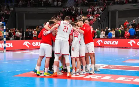 Drużyna piłkarzy ręcznych w białych strojach z czerwonymi elementami tworzy zgraną grupę na boisku.