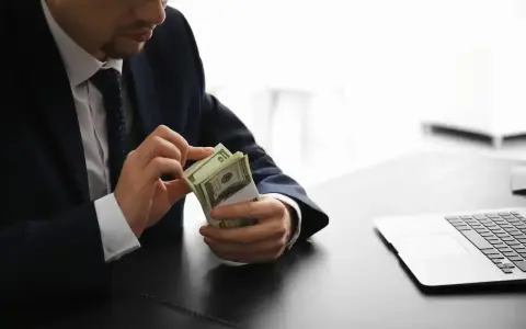 Mężczyzna w garniturze liczy pieniądze, zastanawiając się, ile zarabia osoba pracująca w banku. Obok leży laptop.