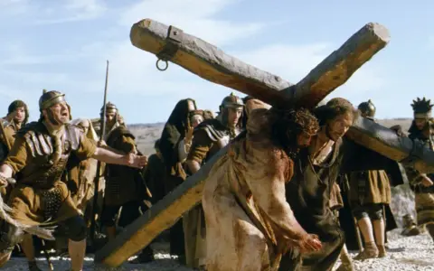 Scena z filmów chrześcijańskich: Jezus dźwigający krzyż, otoczony przez żołnierzy.