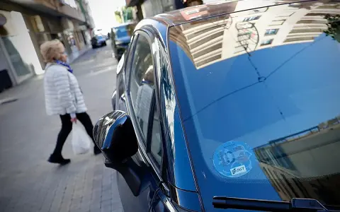 Una mujer cruza la calle mientras un coche eléctrico, con distintivo DGT, espera. Los coches eléctricos pagan zona azul.
