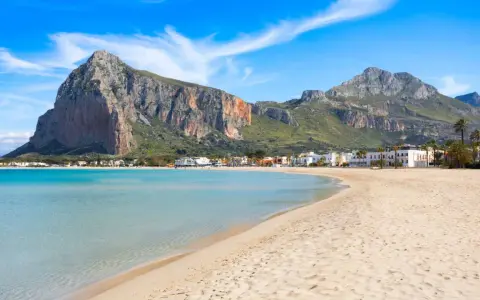 Spiaggia dorata e mare cristallino, un angolo di paradiso in Sicilia dove andare al mare. Montagne imponenti sullo sfondo.