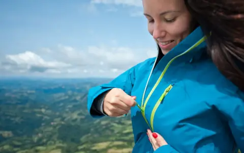Donna con impermeabile montagna blu, chiude la zip. Paesaggio verdeggiante sotto un cielo azzurro.